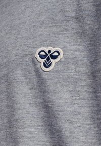 Grå tekstil med et broderet emblem af en bi i blå og hvid farveskema. Stoffet har et blødt, struktureret udseende.