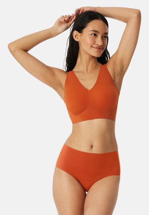 Frau mit langen dunklen Haaren trägt einen nahtlosen orangen BH und passende hoch taillierte Unterwäsche, die Arme über dem Kopf erhoben, lächelnd.