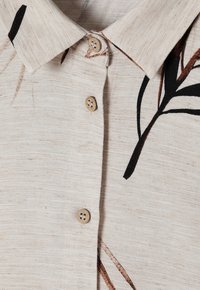 Camisa de un color beige claro con cuello, botones de madera y patrones florales oscuros. Tejido texturizado con detalles de tejido visibles.
