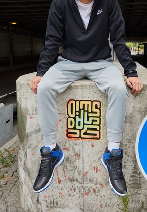 Personne portant des baskets Nike noires et bleues, un pantalon gris clair et une veste noire Nike, assise sur un bloc de béton avec des graffitis colorés.