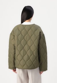 Quilted olivengrønn jakke med en puffy, boble-lignende tekstur, oversized passform og rund hals. Kombinert med lyse bukser.
