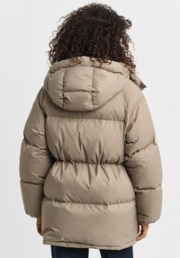 Dikke beige jas met capuchon, met een gequilt ontwerp en elastische manchetten, gezien vanuit de rug. Zachte textuur en duurzaam materiaal.