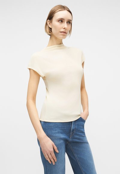 Γυναίκα με κοντά ξανθά μαλλιά, φοράει μπλούζα μπεζ κοντομάνικη και μπλε τζιν, στέκεται με το ένα χέρι στην τσέπη μπροστά σε απλό φόντο.