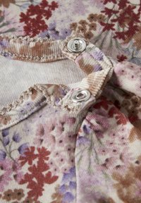 Close-up van stof met bloemenprint in bruin, paars en roodtinten, voorzien van twee zilveren drukknoopsluitingen en decoratieve kartelrandstiksels langs de randen.