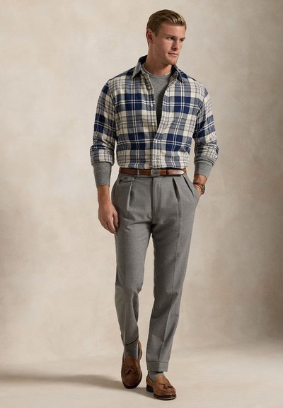 Mand iført en blå og hvid ternet skjorte over en grå sweater, grå plisserede bukser, brun bælte og brune loafers, går med hænderne i lommerne.