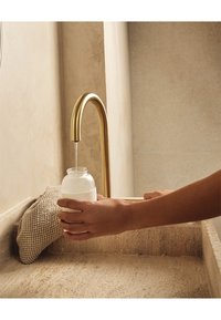 Bottiglia in vetro satinato tenuta sotto un elegante rubinetto dorato, con acqua che scorre sopra un lavandino in pietra beige testurizzata e un accento in tessuto intrecciato.