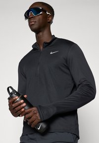 Svart långärmad zip-up tröja med en lätt lyster, med en liten vit Nike-logotyp. Mörkgrå vattenflaska med handtag och metallicelement.