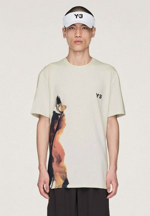 Joven con visera blanca Y-3 y camiseta beige Y-3 con un estampado abstracto en naranja y negro en el lado izquierdo.