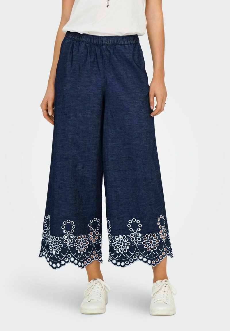 Marineblauwe culottebroek met een elastische tailleband; heeft witte bloemenborduursels aan de zoom. Gemaakt van een textiele stof. Draagt met witte sneakers.