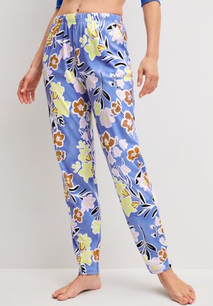 Vrouw met lichtblauwe, hoge taille broek met een bloemenpatroon in geel, bruin, roze en zwart, staand op blote voeten tegen een effen achtergrond.