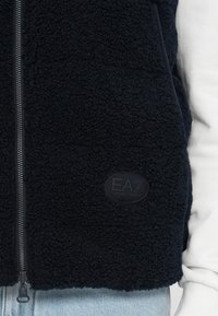 Chandail en polaire marine avec fermeture éclair, tissu texturé et patch logo EA7 Emporio Armani. Un sous-vêtement gris est visible en dessous.
