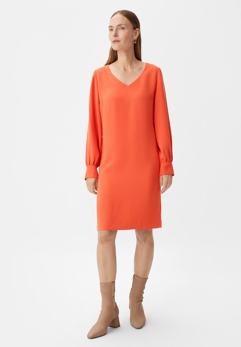 Oranges V-Ausschnitt-Kleid mit langen, gepufften Ärmeln. Weicher, glatter Stoff mit geradem Schnitt, kombiniert mit beigen Ankle Boots.