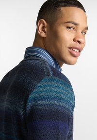 Maglione lavorato a maglia con una superficie testurizzata in tonalità di blu e nero, abbinato a una camicia a righe blu con colletto. Mostra dettagli sulle spalle e vestibilità.