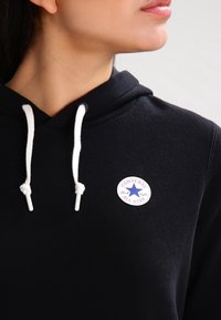 Sweat à capuche noir avec cordon de serrage, doté d'un patch logo en caoutchouc blanc avec une étoile bleue et un texte rouge, fabriqué en tissu doux.