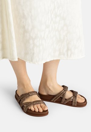 Feet wearing brown glittery braided strap flat sandals with a white textured midi skirt visible above.