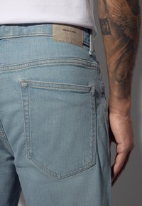 Jean en denim bleu clair avec une finition texturée, doté d'une poche arrière et d'un patch en cuir marqué "DENIM SUPPLY".