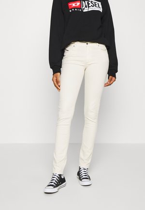 Persoon draagt off-white slim-fit jeans, zwarte Diesel trui met logo en zwarte hoge sneakers met witte veters op een effen achtergrond.