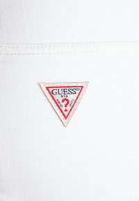 Litet triangulärt Guess-märke med röd kant och text på vit tygbakgrund, som visar sömdetaljer.