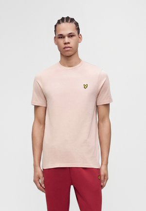 Jeune homme aux cheveux tressés portant un t-shirt rose clair à manches courtes et un pantalon rouge, debout devant un fond uni.