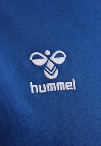 Blått tyg med en vit broderad logotyp med en symmetrisk design och ordet "hummel" nedanför. Texturen är jämn och slät.