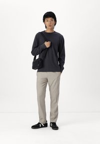 Chemise à manches longues gris foncé, pantalon beige clair et baskets noires avec des rayures blanches. Un petit sac à bandoulière noir pend à l'épaule.