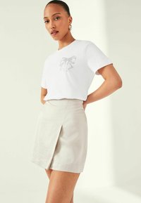 T-shirt blanc à manches courtes avec un graphique de nœud argenté sur la poitrine, associé à une jupe enveloppante beige clair dotée d'une fente sur le côté.