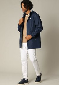 Veste imperméable navy avec fermeture éclair, capuche ajustable et poignets élastiques, associée à un haut beige clair et un pantalon blanc. Baskets décontractées.
