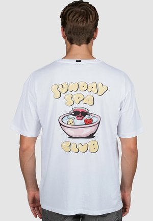 Persona con camisa blanca que lleva un dibujo de un tazón de frutas y agua con texto "Sunday Spa Club" en la parte de atrás, sobre un fondo liso.