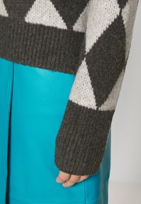 Maglione lavorato a maglia con un motivo a rombi in grigio scuro e crema. La manica presenta un polsino a coste. Sotto, una gonna in pelle turchese brillante.