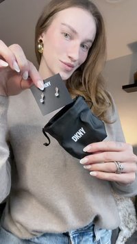 Črni DKNY ovitek vsebuje srebrne uhane v obliki kapljic, prikazane na črni kartici, s dvema okroglima biseroma. Modelov pulover je bež, vidni pa so tudi deli kavbojk.