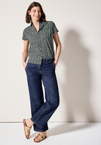 Camicia a maniche corte con motivo verde e nero, abbinata a jeans a gamba larga blu. Il modello indossa sandali intrecciati color beige. Sfondo neutro.