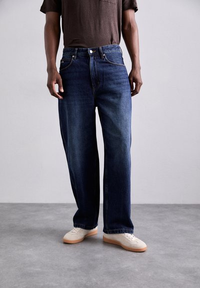 Jeans de mezclilla en azul oscuro, de corte relajado con diseño de pierna recta, con bolsillos frontales y cierre de botón. Llevados con zapatillas de color claro.