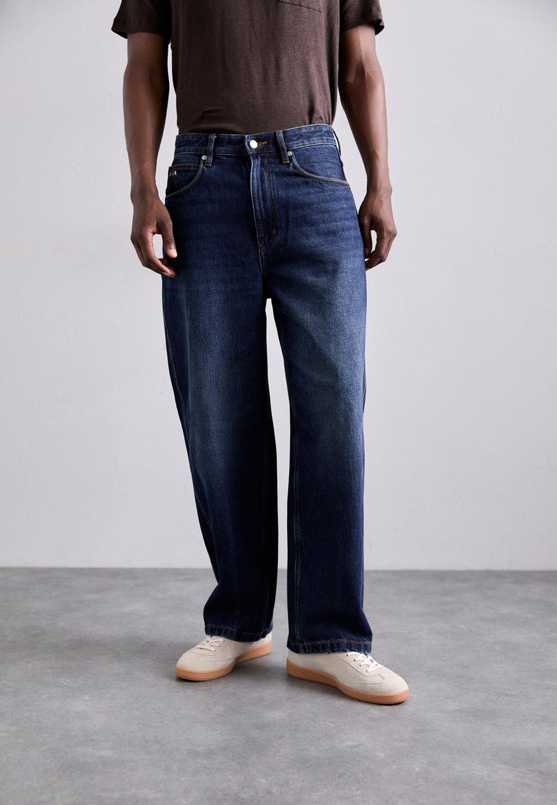 Denimjeans i mörkblått, avslappnad passform med rak bendesign, med framfickor och knäppning. Bärs med ljusa sneakers.