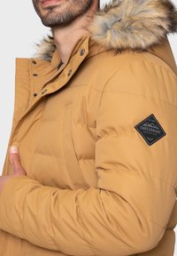 Veste matelassée couleur camel avec capuche bordée de fourrure, texture matelassée, poches avant et un patch logo noir sur la manche.