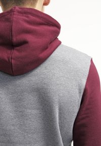 Person som bär en grå huvtröja med vinröda ärmar och huva, sedd bakifrån mot en vit bakgrund.