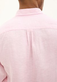 Camicia di lino rosa chiaro con piega sul retro e colletto standard, che mette in mostra un tessuto testurizzato e una vestibilità rilassata.