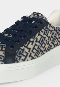 Baskets basses avec lacets bleu marine et motif géométrique bleu marine sur tissu beige, semelle blanche matelassée et col rembourré.