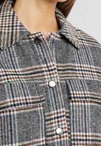 Koszula w kratę w odcieniach szarości, brązu i niebieskiego, z kołnierzem typu point, zatrzaskami i kieszenią na piersi. Tkana tkanina o teksturowanym wykończeniu.