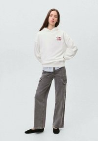 Felpa bianca con testo rosso, abbinata a pantaloni cargo grigi. L'outfit presenta una vestibilità rilassata e texture morbide, con scarpe nere piatte.