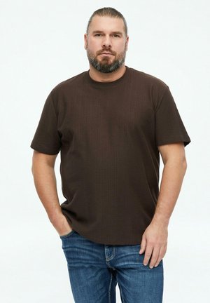 Man met baard en vastgebonden haar, gekleed in een donkerbruin shirt met korte mouwen en blauwe spijkerbroek, staat met één hand in de zak tegen een witte achtergrond.