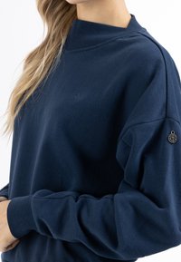 Sweat-shirt bleu marine à col montant, manches longues et texture douce. Comprend un petit logo et un accent métallique décoratif sur la manche.