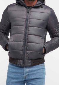 Grå puffjacka med dragkedja, huva och ribbad manschett. Har horisontell sömnad och en logotyp på bröstet. Bärs över blå jeans.