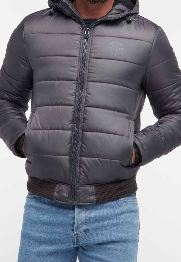 Grå puffjacka med dragkedja, huva och ribbad manschett. Har horisontell sömnad och en logotyp på bröstet. Bärs över blå jeans.