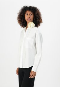 Camicia bianca con bottoni, dotata di una singola tasca sul petto, maniche lunghe e un delicato accento di fiore giallo chiaro sul colletto. Tessuto morbido, vestibilità su misura.