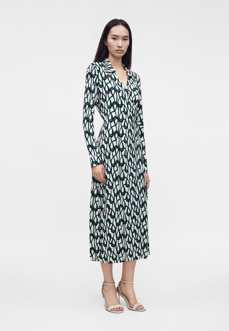 Woman standing wearing a long-sleeve, patterned midi dress with black, white, and teal abstract chain motifs and beige high-heeled sandals.