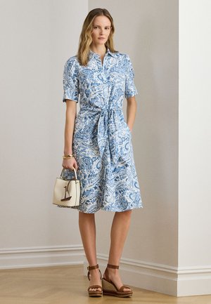 Žena v modrobílých šatech s krátkými rukávy ke kolenům a s uzlem vpředu, držící bílou kabelku s přívěskem lodě.