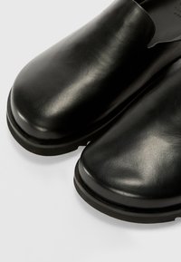 Crne kožne cipele s glatkom teksturom, okruglim vrhom i minimalističkim dizajnom. Ravni gumeni potplat, bez ukrasnih elemenata.