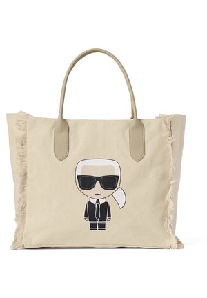 Beige tote tas met franjes aan de randen en twee handvatten, met een cartoonfiguur die een zwart zonnebril, een pak en een witte paardenstaart draagt.