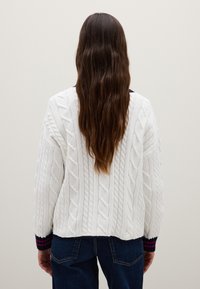 Weißer Kabelstrickpullover mit langen Ärmeln und verkürztem Saum. Gerippte Bündchen mit dunkelblauen und burgunderfarbenen Streifen. Getragen mit dunklen Jeans.