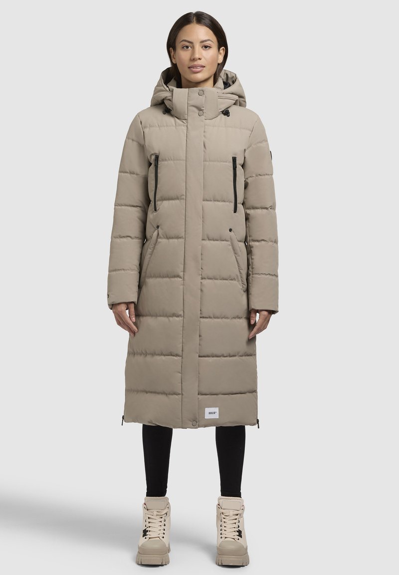 Lang, beige pufferjakke med horisontale quiltede mønster, høj krave, hætte og sorte lynlåslommer. Kombineret med beige støvler og leggings.